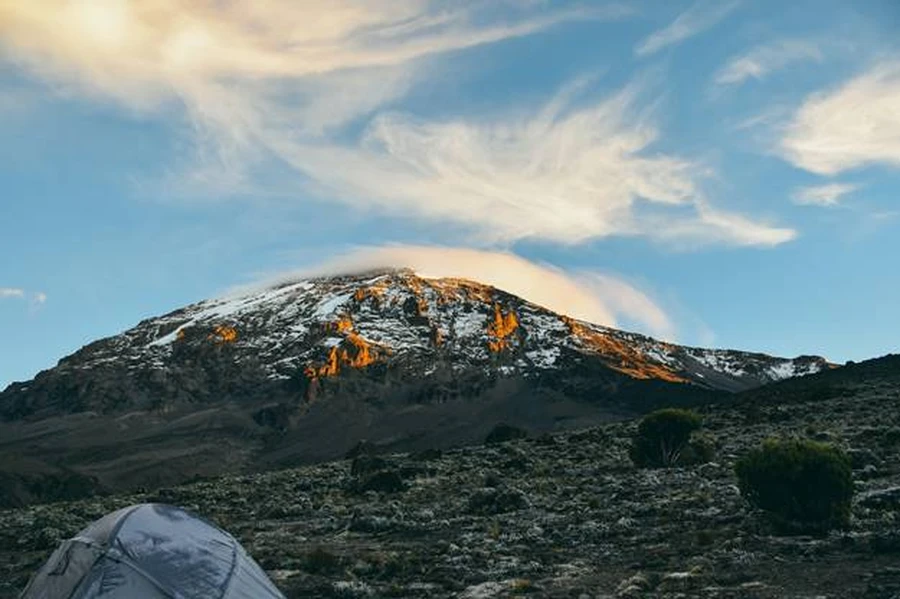 A camp tent set below Mount Kilimanjaro snow-capped peak bathed in golden light of golden hour.