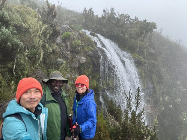 Two hikers in blue rain jackets taking a selfie with their guide in green rain jacket by a waterfall
