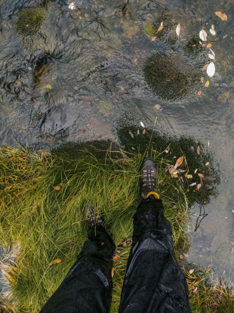 Legs of a hiker near a stream wearing waterproof pants and hiking boots. Looks like it rained. 