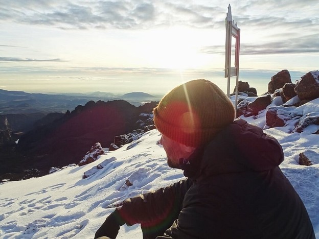Hiker sitting on snowcapped Pt. Lenana enjoying the sunrise wearing warm jacket, beanie, and gloves