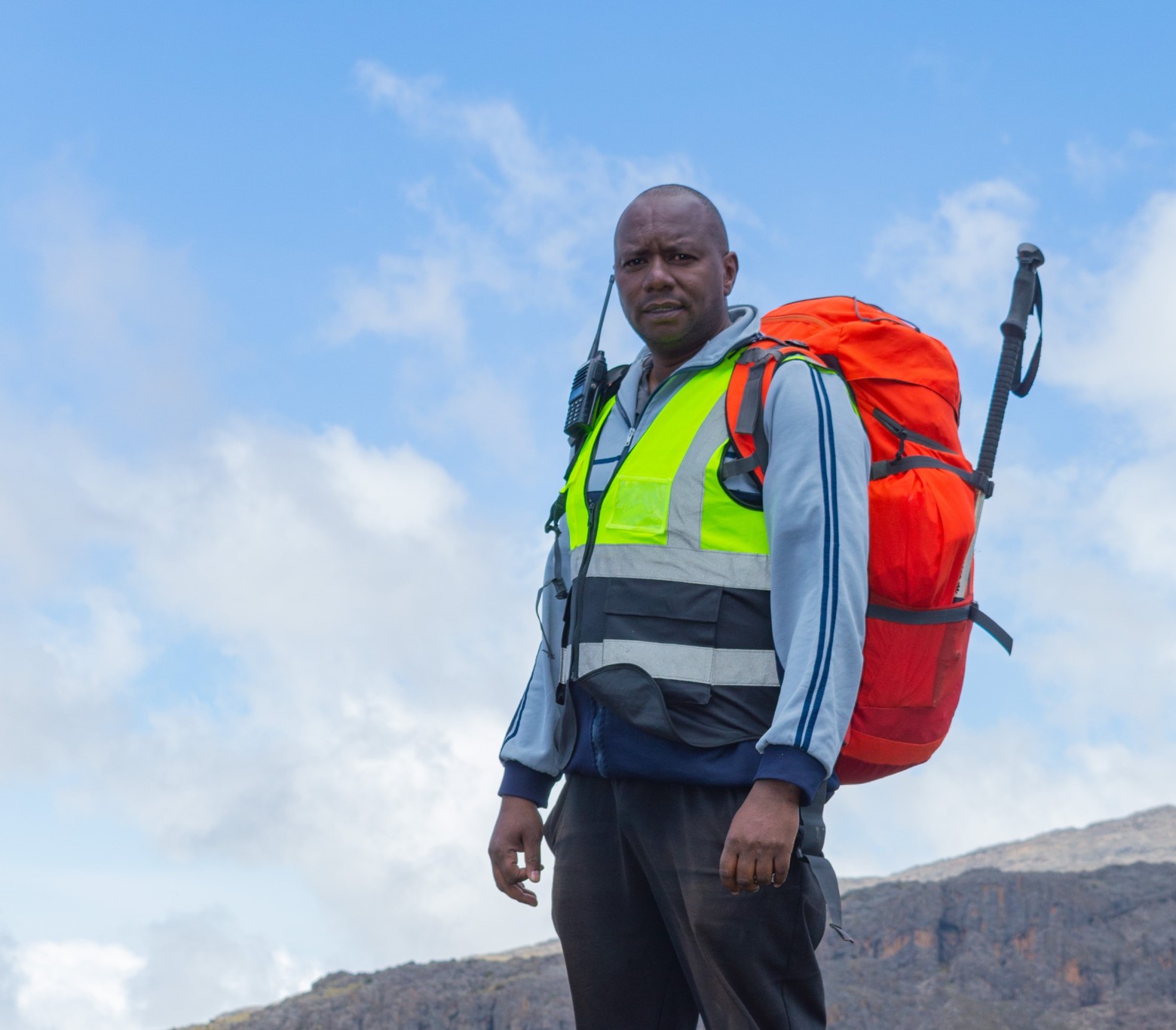A Mount Kenya guide in a reflector jacket, carrying a backpack, a trekking pole and a handheld radio