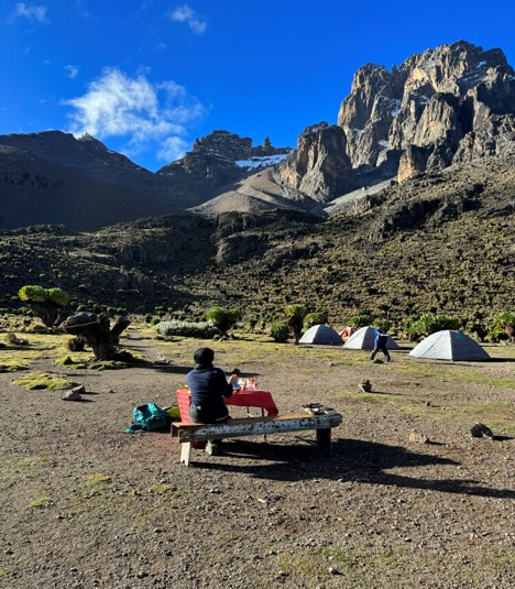 A hiker sitting on a bench having a meal below Mount Kenya peaks at Mackinder’s campsite